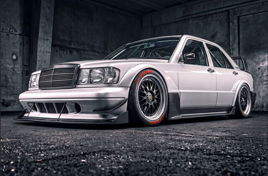 A heavily customized white Mercedes-Benz sedan with a widebody kit, custom front and rear bumpers, and multi-spoke wheels with red accents, parked inside a dark concrete garage.
