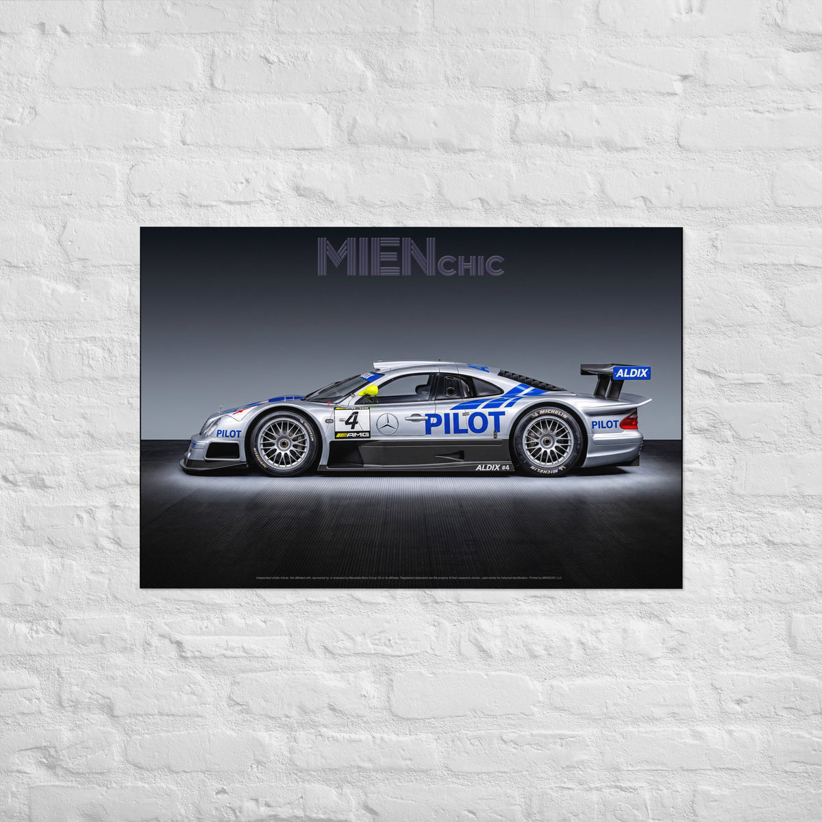 A framed or mounted wall art print showing a side-profile racing car on a dark studio background, hung on a white painted brick wall. The print includes visible text such as MIENCHIC and ALOX branding on the car.